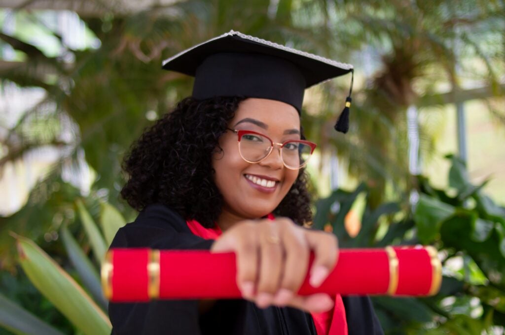 Woman in a black graduation cap and gown with red accents smiling outside